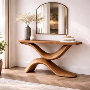 Puede incluir: Una consola de madera con una base curva única y una parte superior rectangular. Un espejo arqueado con marco dorado cuelga sobre la mesa, que está decorada con un jarrón de flores, un candelabro y libros. La mesa es de madera marrón claro.