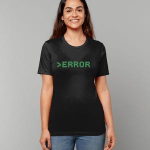 May include: Black t-shirt with the word ">ERROR" in green, pixelated font. The shirt is a crew neck style and is being worn by a person. The person is wearing blue jeans.
