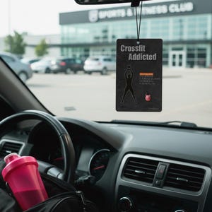 Könnte beinhalten: Ein schwarzer Auto-Lufterfrischer mit dem Text "Crossfit Addicted" und einer Grafik einer Person, die Gewichte hebt. Eine rosa Wasserflasche befindet sich in der Konsole des Autos. Der Hintergrund zeigt einen Sportverein und geparkte Autos.
