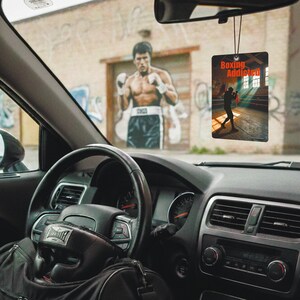 Könnte beinhalten: Ein rechteckiger Auto-Lufterfrischer mit den Worten "Boxing Addicted" in Rot. Das Bild auf dem Lufterfrischer zeigt einen Boxer in einem Ring. Der Lufterfrischer hängt am Rückspiegel in einem Auto. Im Hintergrund ist ein Wandbild eines Boxers zu sehen.