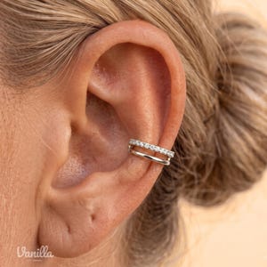 Könnte beinhalten: Ein silberfarbener Ohrring in Form einer Manschette mit zwei parallelen Bändern. Das obere Band ist mit einer Reihe kleiner, klarer, funkelnder Steine besetzt, während das untere Band aus glattem, poliertem Metall besteht. Am Knorpel platziert.
