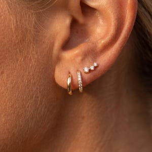 Könnte beinhalten: Ein Set aus drei goldenen Ohrringen am Ohrläppchen. Das Set besteht aus einem schlichten, polierten Goldring, einem mit kleinen klaren Steinen besetzten Goldring und einem goldenen Ohrstecker mit drei kleinen klaren Steinen in einer Reihe.