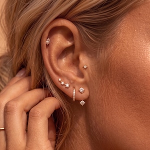 May include: A collection of gold-toned earrings worn in a single ear, featuring a marquise-cut stud, a small star-shaped stud, a three-stone curved climber, a small hoop, and two round brilliant-cut solitaire studs with clear stones.