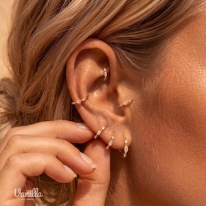 Könnte beinhalten: Ein Set aus fünf goldenen Creolen, die in einem Ohr getragen werden, jede mit kleinen, klaren, marquise-geschliffenen Steinen entlang des Rings. Die Ohrringe sind im Rook-, Conch- und Ohrläppchen-Piercing platziert und aus goldfarbenem Metall.