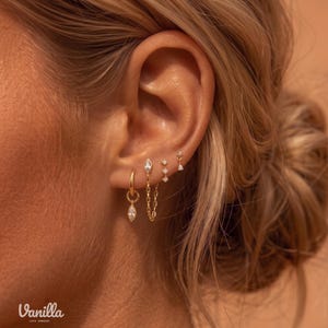 Op de afbeelding: Een set goudkleurige oorbellen bestaande uit een kleine ring met een marquise-geslepen steenbedel, een kettingoorbel met een marquise-geslepen steen, een verticale knop met drie stenen en een kleine driehoekige steenknop. Tekst Vanilla 1978 Jewelry zichtbaar.