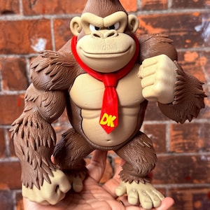 May include: A brown gorilla figurine with a tan chest and face, wearing a bright red necktie featuring the yellow letters DK. The figure has a muscular build, textured fur detailing, and a determined facial expression with visible teeth.