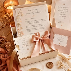 Puede incluir: Una caja de regalo que contiene papelería con el texto "OPEN WHEN... you need to feel close to me" y "OPEN WHEN LETTERS FOR MY HUSBAND". El conjunto incluye una cinta de satén rosa empolvado, un sobre a juego con la etiqueta "You Need To Feel Loved" y una tarjeta con una cita sobre sentirse seguro en el mundo, todo decorado con ilustraciones florales.