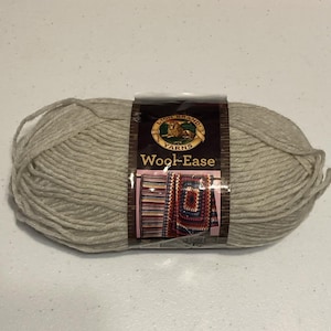 May include: A large ball of light beige Lion Brand Wool-Ease yarn. The label features the brand name, a lion logo, and images of knitted projects. The yarn is tightly wound and ready for knitting or crocheting.