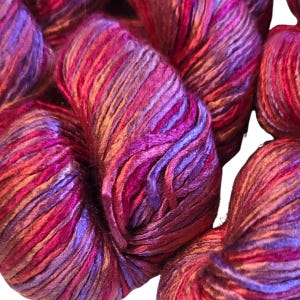 May include: Close-up of several skeins of yarn in shades of purple, red, and orange. The yarn has a textured appearance, with visible strands and a slight sheen. The colors blend together, creating a vibrant and rich visual effect, suitable for knitting or crochet projects.