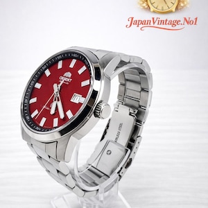 May include: A stainless steel Orient watch with a vibrant red dial and a date window at the 3 o'clock position. The watch features a silver bezel with black markings and a matching stainless steel band. The text "Orient" is visible on the dial. The watch is displayed on a clear stand.