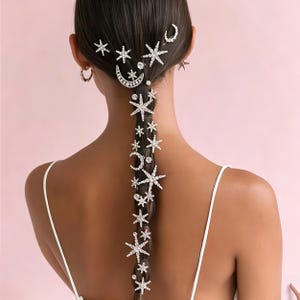 May include: A set of silver-toned hair accessories featuring multiple star and crescent moon shapes encrusted with clear rhinestones. These decorative pieces are arranged along a long, dark braided hairstyle, creating a celestial-themed pattern.