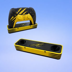 Puede incluir: Un mando de juego negro y amarillo con un patrón de marcas de garras, apoyado sobre un soporte rectangular a juego. Un segundo soporte idéntico se sitúa debajo, mostrando dos aberturas circulares y un diseño de garras en la parte superior.