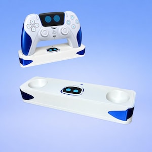 May include: A white and blue gaming controller charging dock featuring a central black display with two blue circular icons. The dock has two circular recessed slots for controllers and blue accents on the sides, matching the controller design.