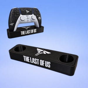 Puede incluir: Un soporte rectangular negro para mando con el texto blanco THE LAST OF US y un logotipo de polilla blanco. Un soporte se muestra vacío, mientras que otro sostiene un mando de juego blanco y negro con gráficos personalizados y el mismo logo.