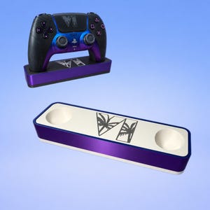 Puede incluir: Un mando de videojuegos inalámbrico con acabado degradado en azul y morado, con un logotipo de palmera y el texto VI, sobre una base de carga morada a juego. Debajo, una segunda base de carga rectangular con la parte superior de color blanco.