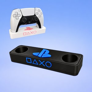 May include: A white gaming controller resting on a white rectangular stand with pink PlayStation symbols, and a separate black rectangular stand featuring two circular cutouts, a blue PlayStation logo, and blue square, triangle, X, and circle symbols.