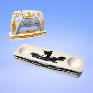 Pode incluir: Um comando de jogo branco com padrões de pinceladas douradas, pousado num suporte a condizer branco e dourado. Por baixo, um suporte retangular branco separado com duas reentrâncias circulares e um design gráfico preto.