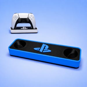 Puede incluir: Un mando de juego blanco y negro descansa sobre un soporte blanco rectangular con un logotipo de PlayStation. En primer plano, un soporte largo y rectangular azul y negro presenta dos aberturas circulares y un logotipo azul de PlayStation.
