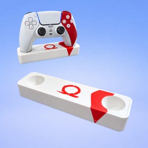 Könnte beinhalten: Ein weiß-roter Gaming-Controller-Ständer mit einem roten Omega-Symbol. Der Ständer verfügt über zwei kreisförmige Aussparungen zur Aufnahme von Controllern, wobei ein weiß-roter Controller im linken Steckplatz der Basis eingesetzt ist.