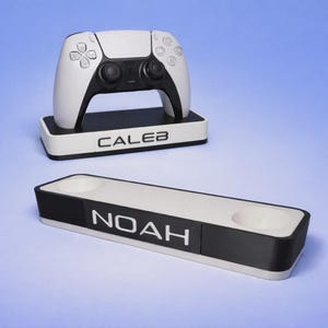 May include: A white and black gaming controller stand featuring the name CALEB in black lettering on the front. Below it, a separate white and black rectangular stand with two circular cutouts features the name NOAH in white lettering.