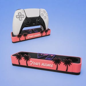 May include: A rectangular controller stand with a pink base, black top, and black palm tree silhouettes. The front features the white text "Your name" and a purple geometric emblem. One stand holds a white and black game controller, while another is empty.
