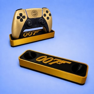 Puede incluir: Un mando de juego inalámbrico dorado y negro con el logotipo 007 en el panel táctil, apoyado sobre un soporte de exhibición rectangular a juego en dorado y negro con el logotipo 007, acompañado de un segundo soporte de exhibición vacío.