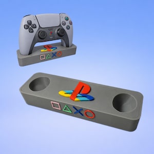 May include: A grey controller stand featuring a colourful PlayStation logo and the iconic square, triangle, cross, and circle symbols on the front. The stand includes two circular cutouts for holding controllers, shown with a grey game controller.