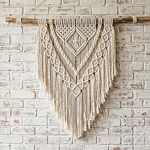 Puede incluir: Tapiz de macramé color crema con un diseño geométrico. La pieza está suspendida de un listón de madera natural. El macramé presenta patrones de diamantes y galones, con largas franjas en la parte inferior. El fondo es una pared de ladrillo blanco.