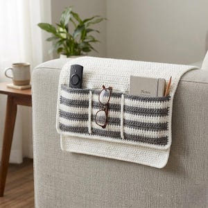 May include: A white crocheted sofa arm organizer with gray and white striped pockets. The pockets hold a remote control, eyeglasses, a notebook labeled "Notes", and a pencil. The organizer is draped over the arm of a gray sofa.