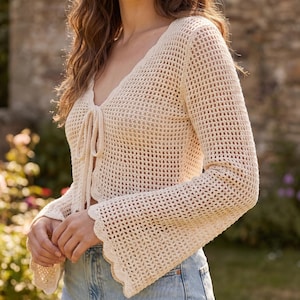 May include: A cream-colored, long-sleeved crochet cardigan featuring an open-knit pattern, a V-neckline, and a front tie closure. The garment has flared sleeves with a scalloped edge finish, paired with light blue denim jeans.