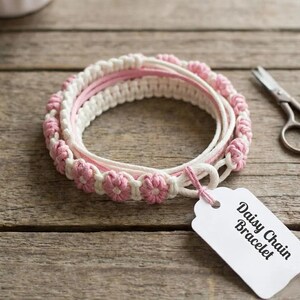 Pode incluir: Uma pulseira enrolada feita de cordão branco com pequenos nós em forma de flor rosa em todo o design. Uma etiqueta retangular branca presa à pulseira exibe o texto impresso em preto Daisy Chain Bracelet em uma fonte sem serifa.