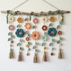 Puede incluir: Un tapiz de pared de ganchillo hecho a mano con flores y hojas coloridas suspendidas de una rama de madera natural. Las flores son en tonos de rosa, azul, amarillo y naranja, con hojas y borlas verdes.