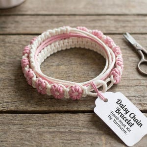 Puede incluir: Un kit de pulsera de cadena de margaritas hecha a mano con cordón de macramé blanco y rosa. La pulsera presenta un diseño de flores de margarita y está etiquetada como "Daisy Chain Bracelet Hand-Knot Kit by Yarnivora."