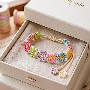 May include: A handmade floral bracelet with crocheted flowers in shades of pink, yellow, green, and blue. The bracelet is displayed in a white gift box, which has the text "Handmade Floral Co."