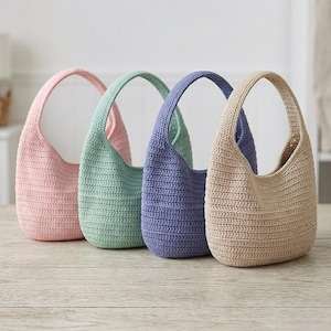 May include: Four crochet shoulder bags in pastel colors: pink, mint green, periwinkle, and beige. Each bag has a rounded shape with a single handle. The bags are displayed on a light-colored wooden surface.