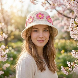 Peut inclure: Un chapeau cloche en crochet crème orné d'applications de fleurs roses et jaunes. Le chapeau a un large bord et est porté par une personne aux longs cheveux châtain clair. L'arrière-plan présente des fleurs roses et de la verdure floues.