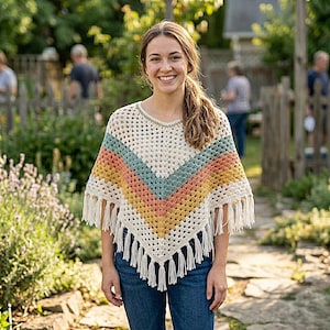 May include: A cream-colored crochet poncho with a colorful chevron design in shades of orange, teal, and yellow. The poncho has fringe along the bottom edge. The garment is worn over a blue denim shirt.