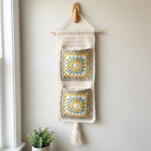 May include: Cream-colored crochet wall organizer with two square pockets featuring a yellow, blue, and cream floral design. The organizer hangs from a wooden dowel and has a cream tassel.