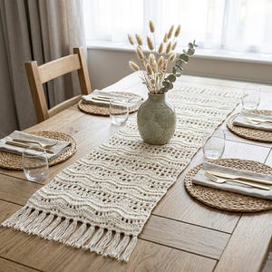 May include: A cream-colored, crocheted table runner with a wavy pattern and fringe detail. The runner is placed on a wooden table, alongside woven placemats, napkins, and glassware. A vase with dried flowers is in the center.