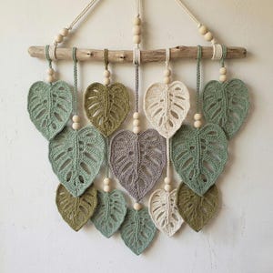May include: A handmade wall hanging with crocheted Monstera leaves in green, cream, and grey. The leaves are suspended from a driftwood branch with wooden beads, creating a natural, bohemian look. This decorative piece adds a touch of nature to any room.