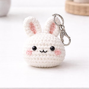 May include: A white crochet bunny keychain with pink inner ears and cheeks. The bunny has black eyes and a stitched mouth. A silver-colored metal clasp is attached to the top of the bunny's head.