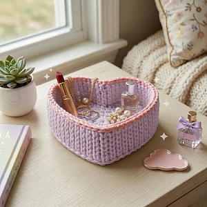 May include: A heart-shaped, lavender-colored, crocheted basket with a pink trim, holding jewelry and perfume bottles. A small succulent in a white pot and a perfume bottle with a purple bow are also on the table. A book and a cloud-shaped dish are also visible.
