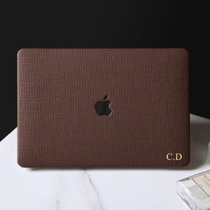 May include: A dark brown textured laptop skin applied to a laptop lid, featuring a cutout for the Apple logo in the center and the gold-colored initials C.D printed in the bottom right corner. The skin has a cross-hatched, leather-like pattern.