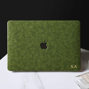 May include: A forest green laptop cover with a textured finish featuring a central cutout for an Apple logo and the gold-colored initials S.A. printed in the bottom right corner. The cover has a rectangular shape with rounded corners.