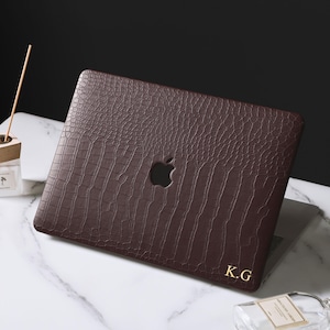 May include: A dark brown laptop skin with a textured crocodile pattern applied to the lid. The skin features a precise cutout for the Apple logo and includes the gold-colored initials K.G printed in the bottom right corner of the cover.