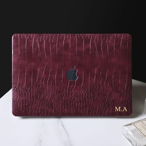 May include: A dark burgundy laptop cover with a textured crocodile skin pattern. The cover features a central cutout for an Apple logo and the gold-colored initials M.A. printed in the bottom right corner, providing a sleek and protective finish.