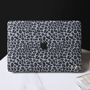May include: A laptop skin featuring a grey and black leopard print pattern with a central cutout for the Apple logo. The bottom right corner of the skin displays the gold-colored initials K.R. in a serif font, providing a personalized detail.