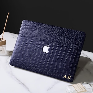 May include: A dark blue laptop cover with a textured crocodile skin pattern. The cover features a central cutout for the Apple logo and the gold-colored initials A.K. printed in the bottom right corner of the rectangular protective shell.