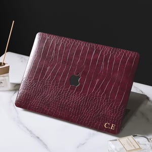 May include: A dark burgundy laptop cover with a textured crocodile skin pattern and the gold embossed initials C.E. in the bottom right corner. The cover features a central cutout revealing the Apple logo on the laptop lid underneath.