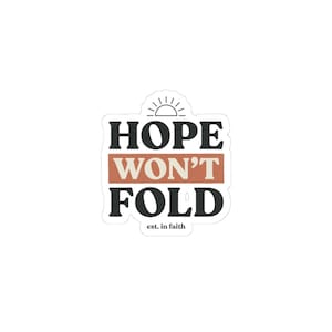 May include: A white sticker with the words "HOPE WON'T FOLD" in black block letters, with a rust-colored horizontal bar between "HOPE" and "WON'T". A small sun graphic is above the word "HOPE". The phrase "est. in faith" is below "FOLD".